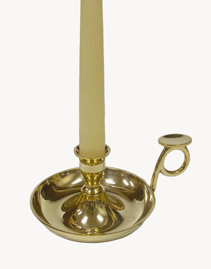 A solid brass chamberstick with a polished, lacquered finish, featuring a flat circular base and a handle for carrying. A candle is placed on the holder.