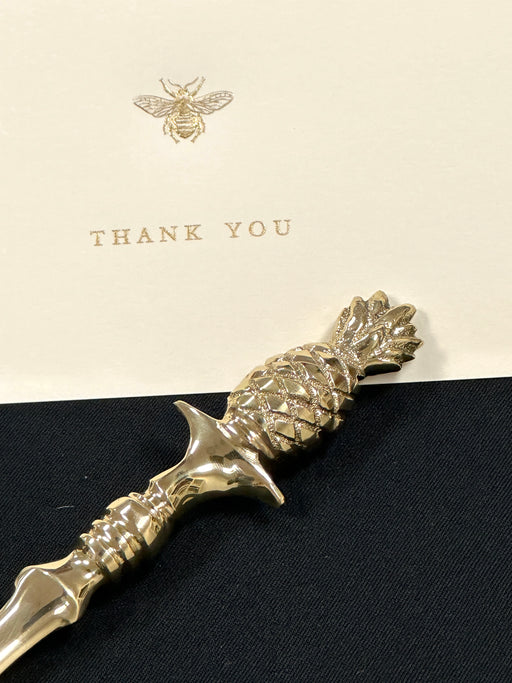 A gold lacquered brass pineapple-shaped letter opener with a detailed handle and an elegantly-shaped blade, placed on a greeting card.