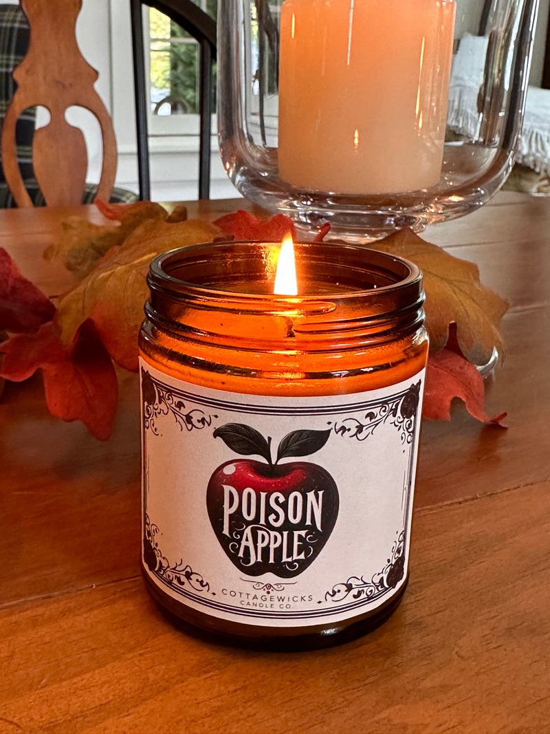 A lit candle in an amber jar with a label that reads 'POISON APPLE', placed on a table with fall leaves around.