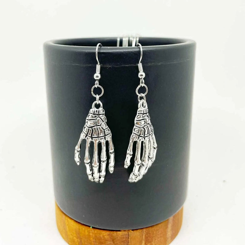 Silver skeleton hand earrings on a black cylindrical stand with wooden base