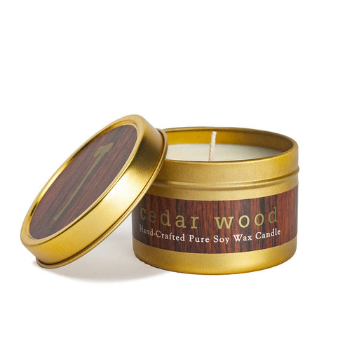 A tin candle with a gold lid and brown label that reads 'Cedar wood'. The candle is shown with the lid off, revealing a white candle inside.