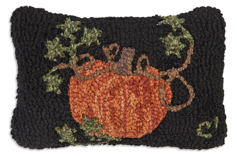 A hand hooked wool pumpkin pillow with a black background featuring a orange pumpkin and green leaves design, made with a velveteen backing and poly-fill pillow insert.